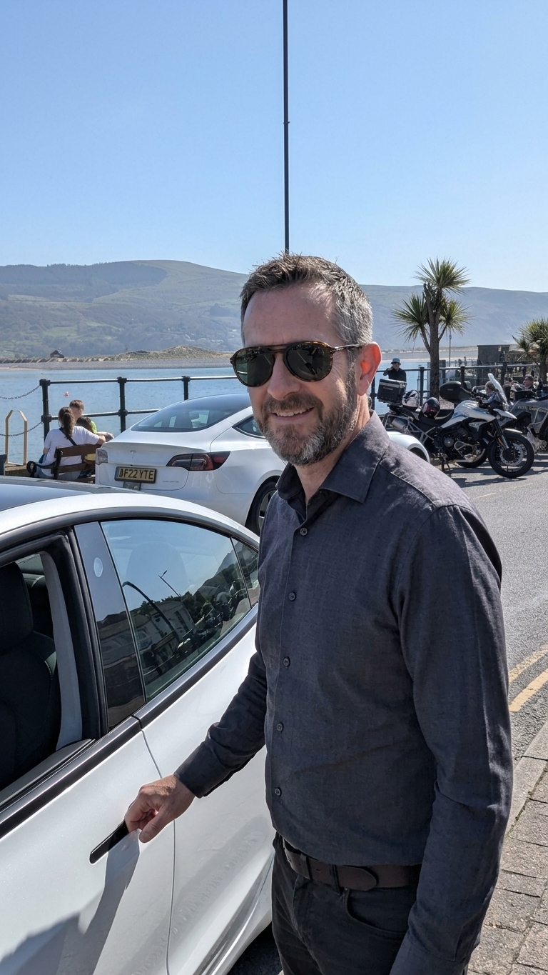 Russell, owner and driver at Gwynedd Airport Taxis, beside his white Tesla Model 3 on the seafront at Barmouth